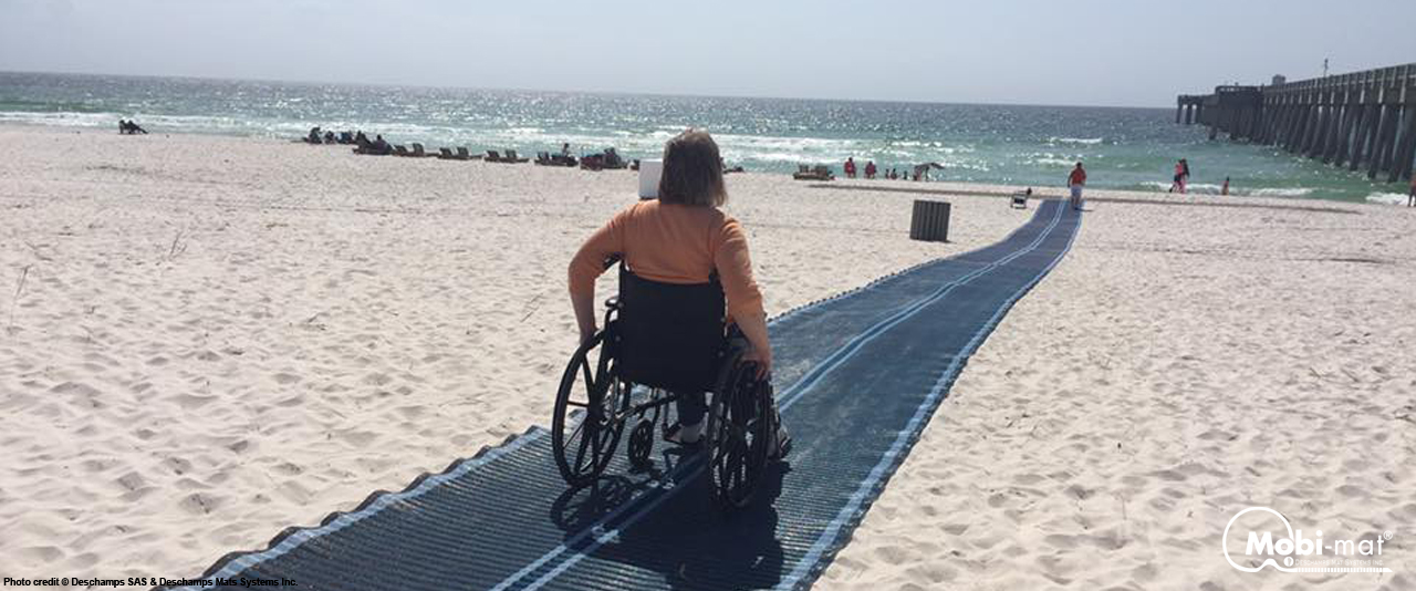The mobi mat is rolled out onto a beach. A woman is using a wheelchair to go to the surf