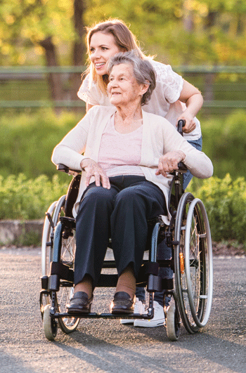 A photo of a young woman pushing an older woman in a wheelchair through a park