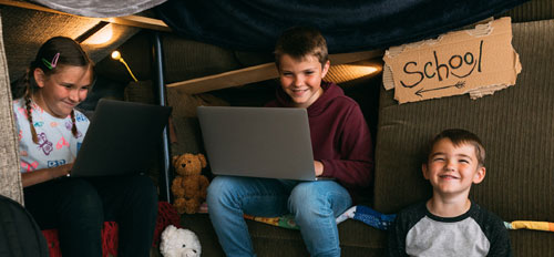 a photo of 3 kids on laptops with a cardboard sign in the background that reads 'School'